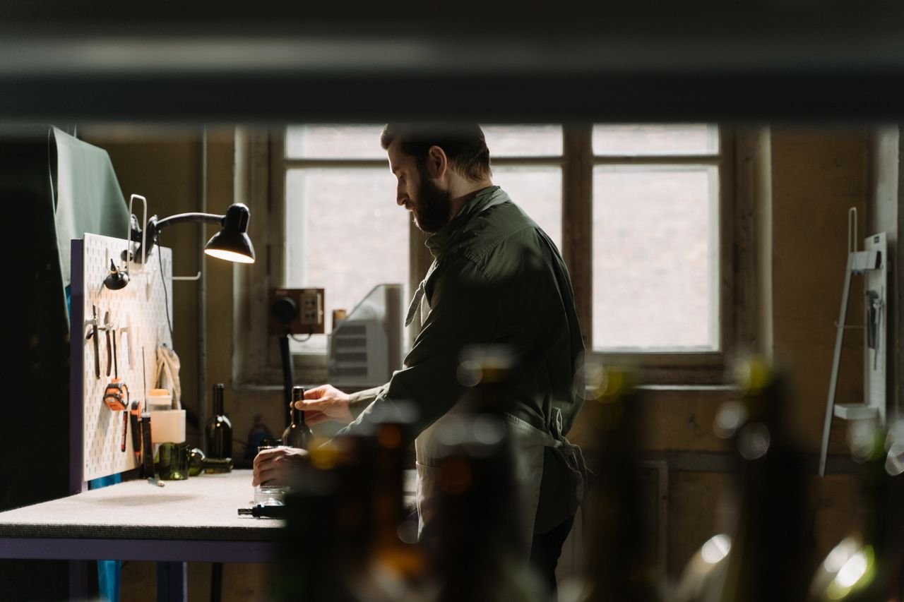Craftsman in workshop with tools and bottles, artistic lighting, and industrial ambiance.